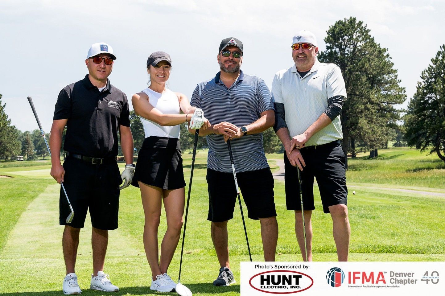Four people pose on a golf course, holding clubs. Sunny day.