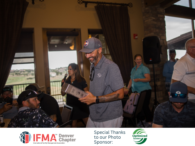 Man in cap holding award, smiling. At IFMA Denver event. People in a room, golf course view.