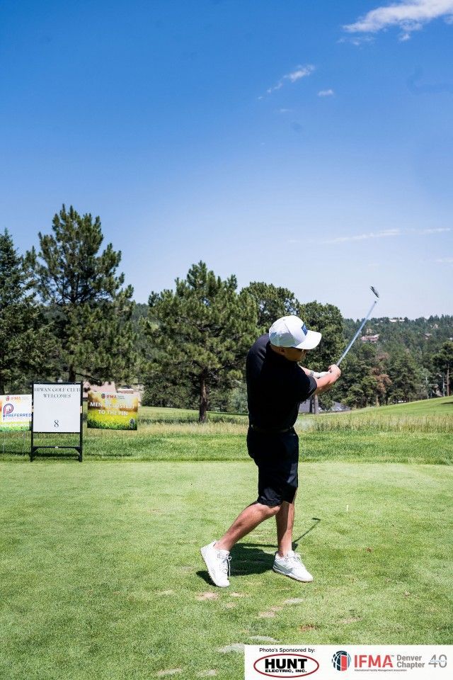 Golfer in black shirt and shorts swinging a club on a sunny green golf course.
