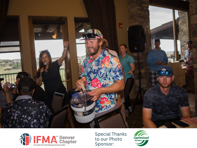 Man in floral shirt holds a bucket, surrounded by people at an event indoors with a scenic view.