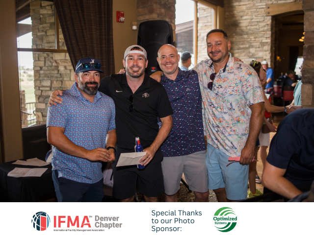 Four men in casual attire pose, arms around each other, indoors at an event.