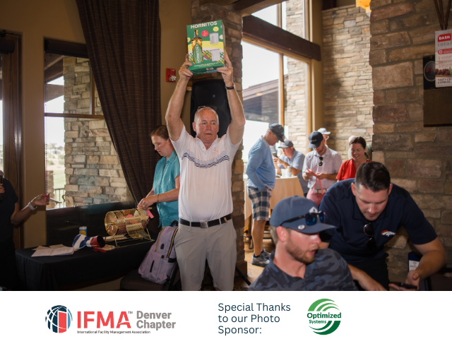 Man holds up a prize at an IFMA Denver Chapter event. People in a room, windows in the background.