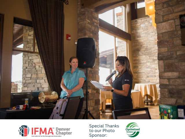Two women at a speaking event: one with a microphone, reading; the other standing nearby. Indoors, near a window.