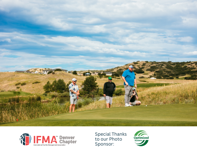 Golfers on a green on a sunny day. One golfer readies a putt, others watch. Hills and a pond in the background.