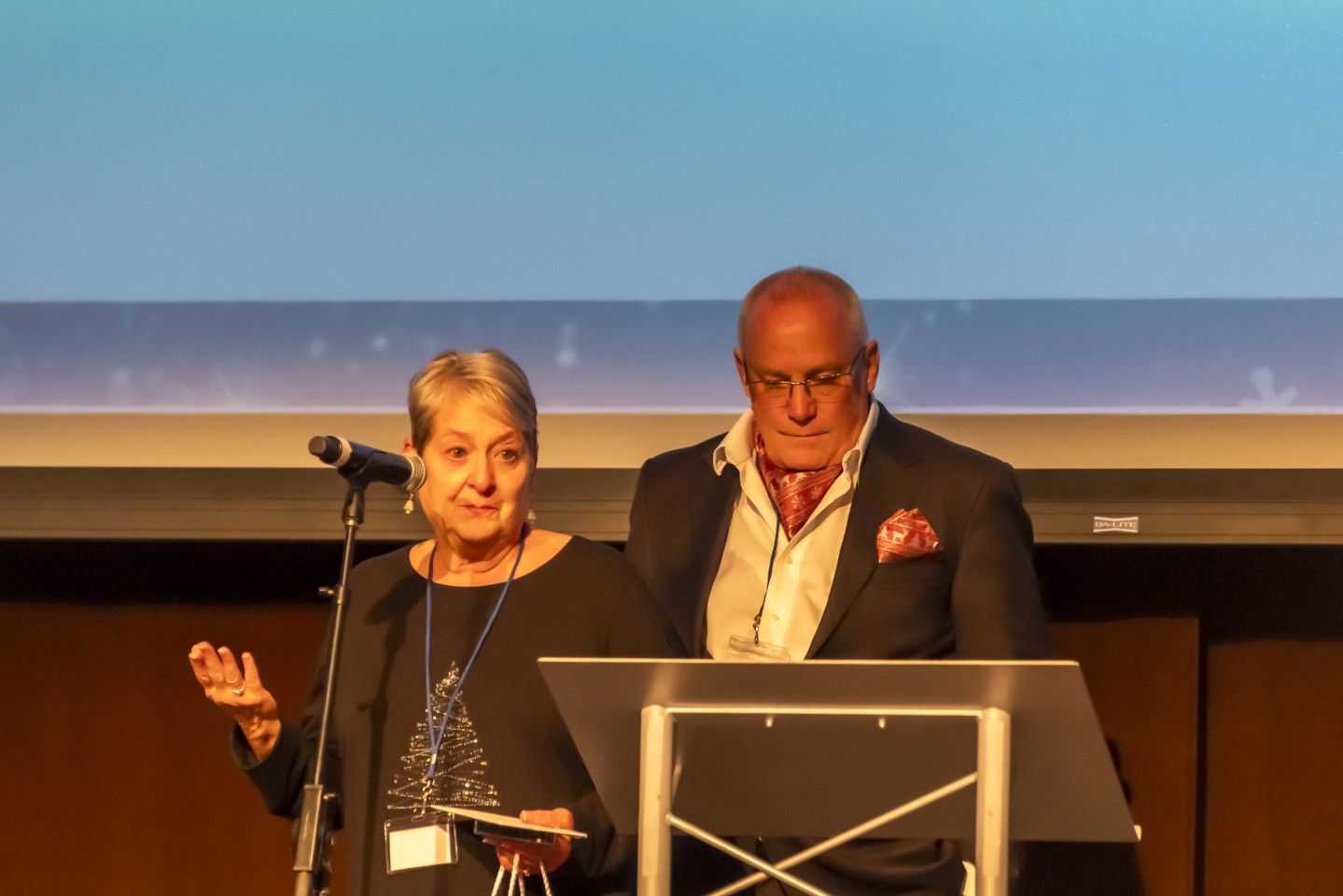 Two people at a podium. Woman on left gestures, man on right looks down. Indoors.
