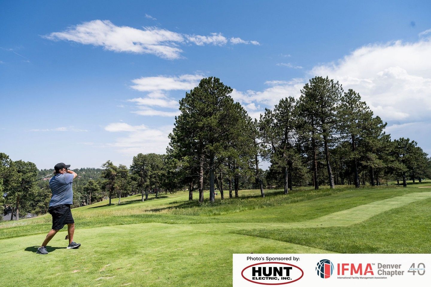 Golfer swinging on a green golf course, trees in the background, under a blue sky.
