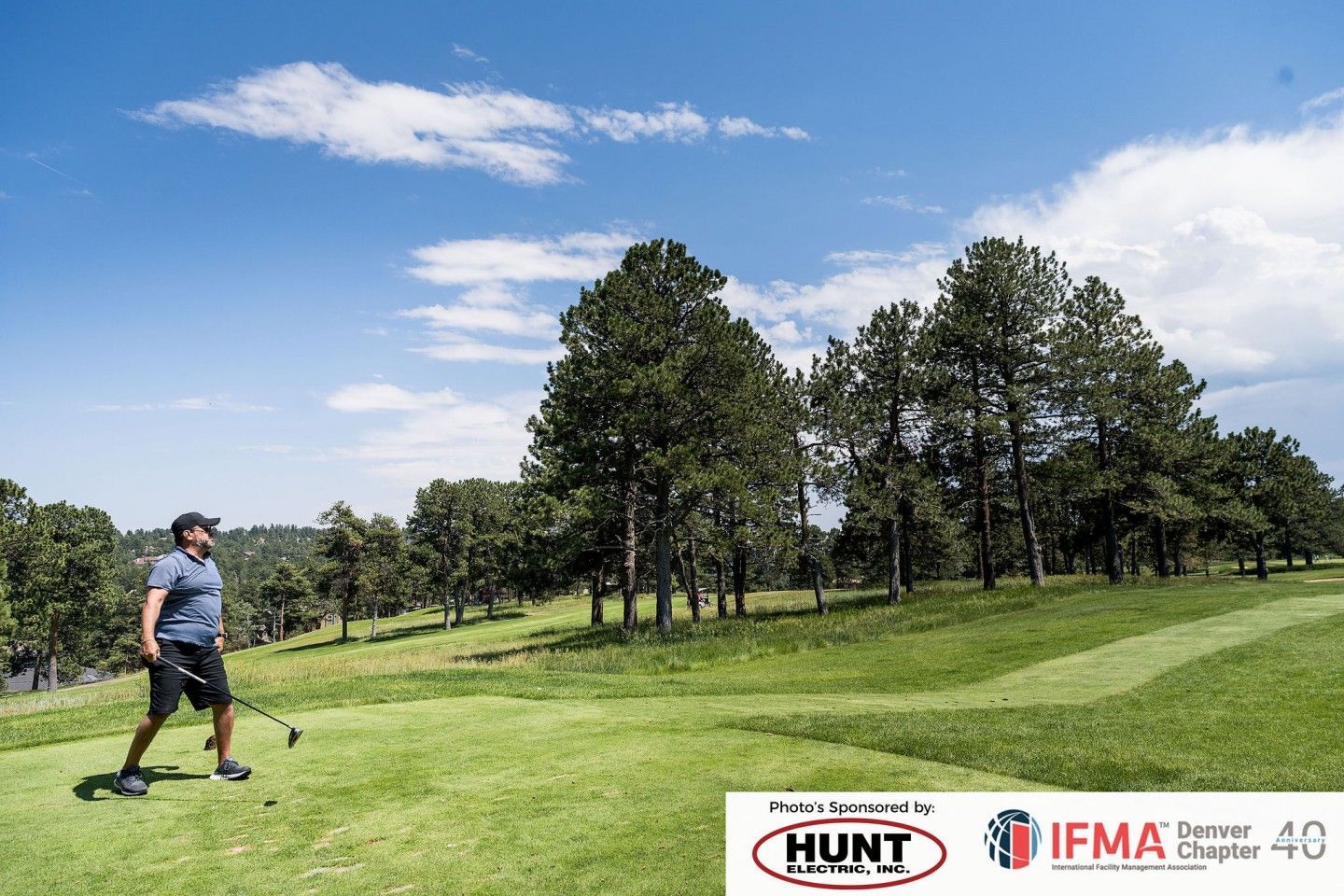 Man swinging a golf club on a green course, with trees and blue sky with clouds.