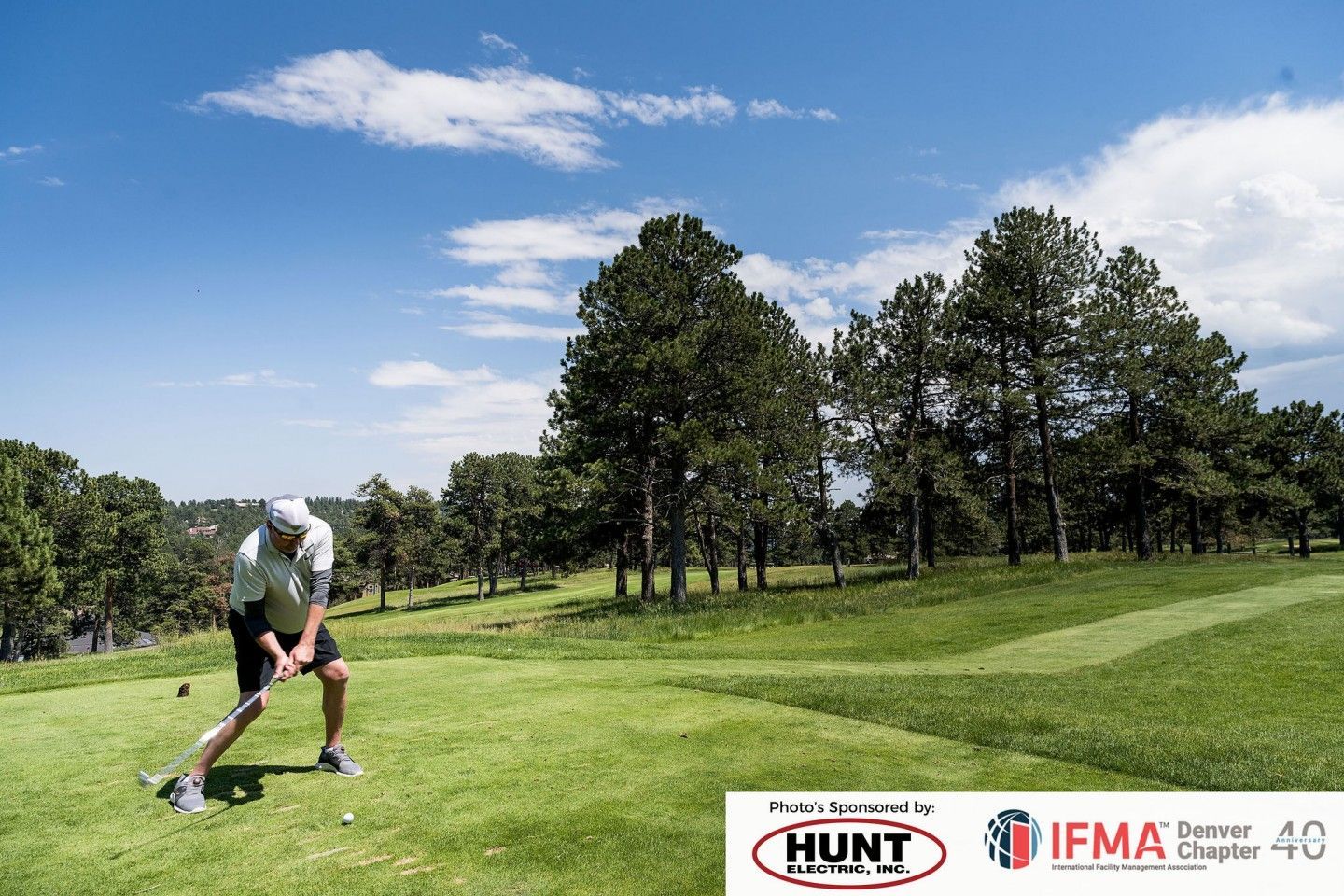 Golfer swings club on green course with trees and blue sky in background.