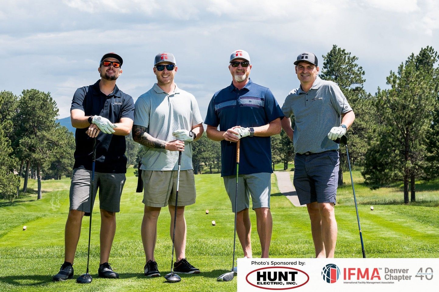 Four men with golf clubs pose on a green, trees and course in the background. Sponsors' logos are at the bottom.