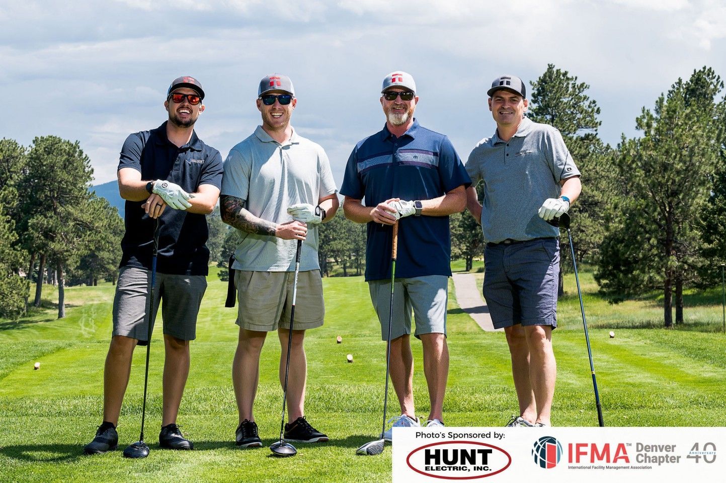 Four men on a golf course holding clubs, smiling, wearing hats and casual clothes.