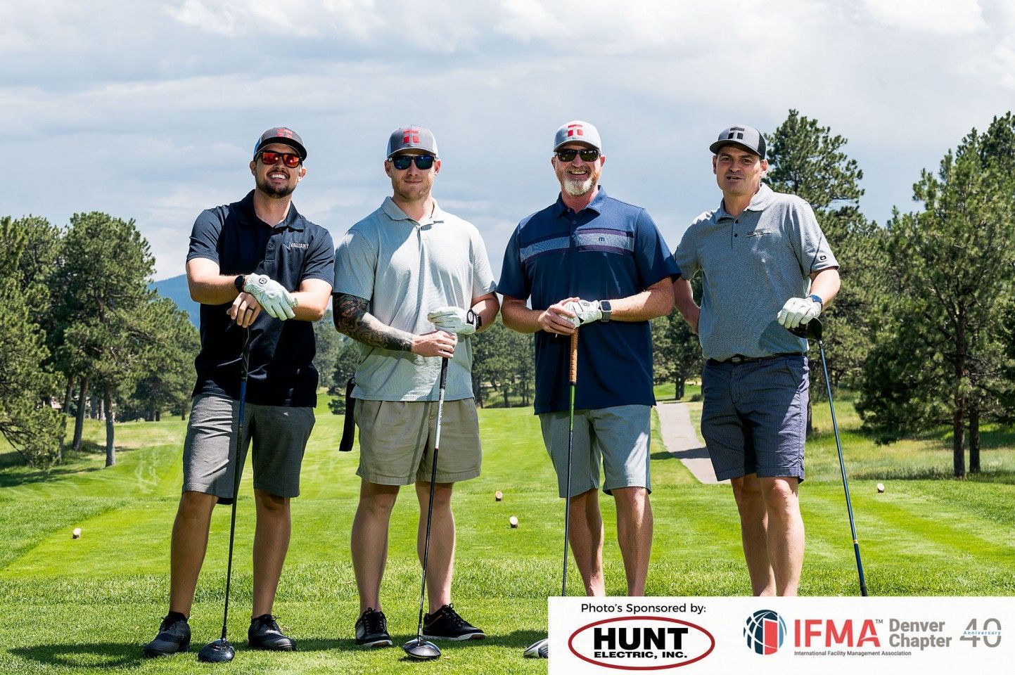 Four golfers stand on a green, holding clubs. Golf course in the background, sunny day.