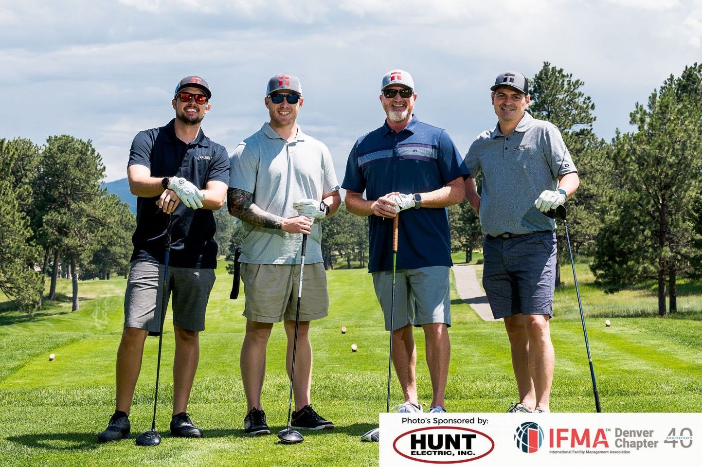 Four men on a golf course holding clubs, smiling. Sunny day, green grass, trees.