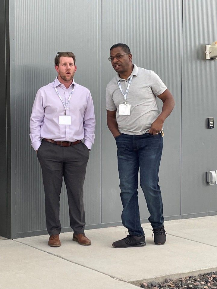 Two men stand outside a building. One wears a light purple shirt and gray pants. The other has on a gray shirt and jeans.