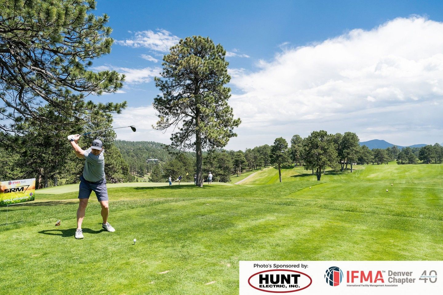 Golfer swings club on green course with trees under a blue sky.