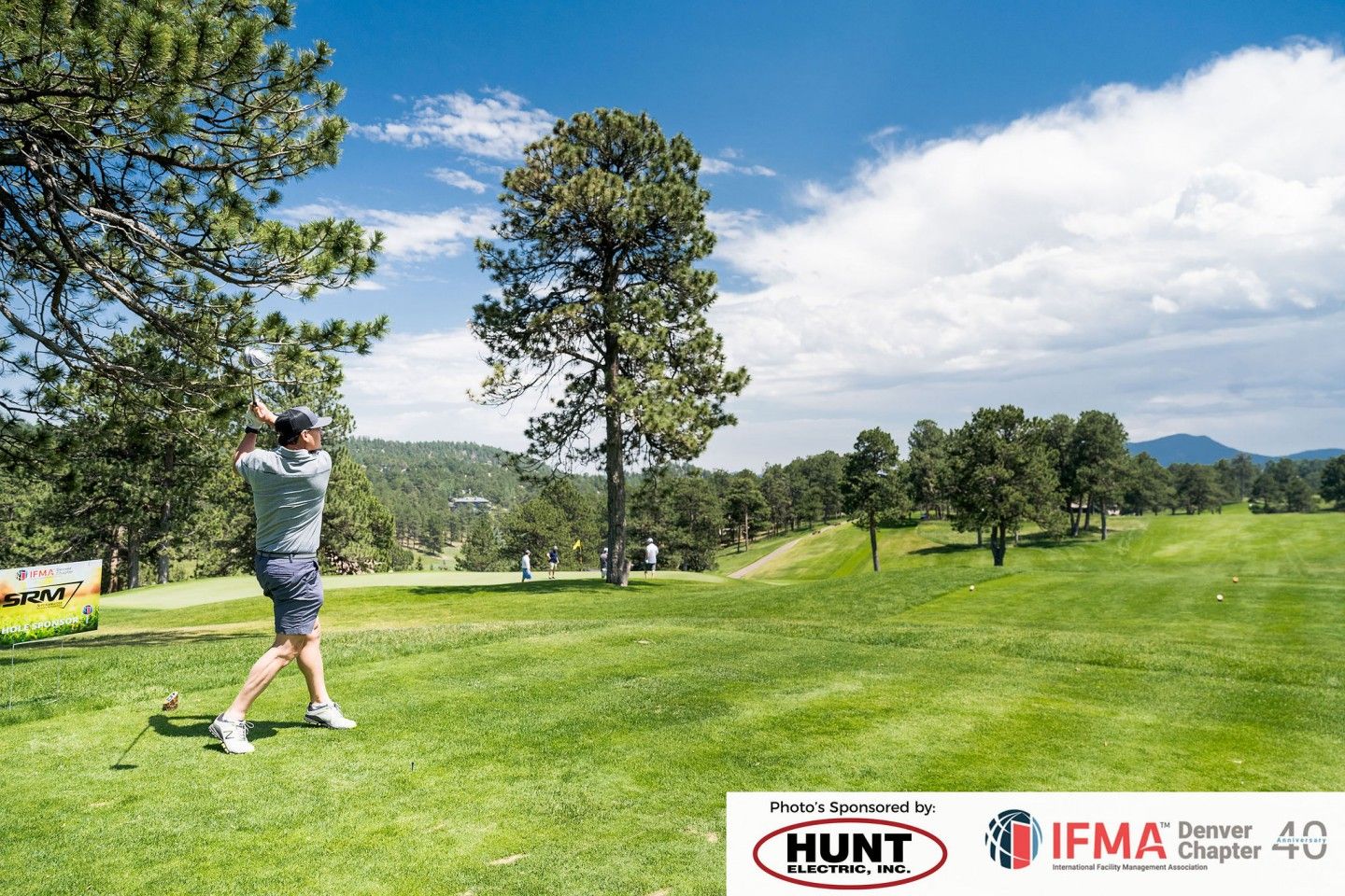 Golfer swings on a green golf course under a blue sky with trees.