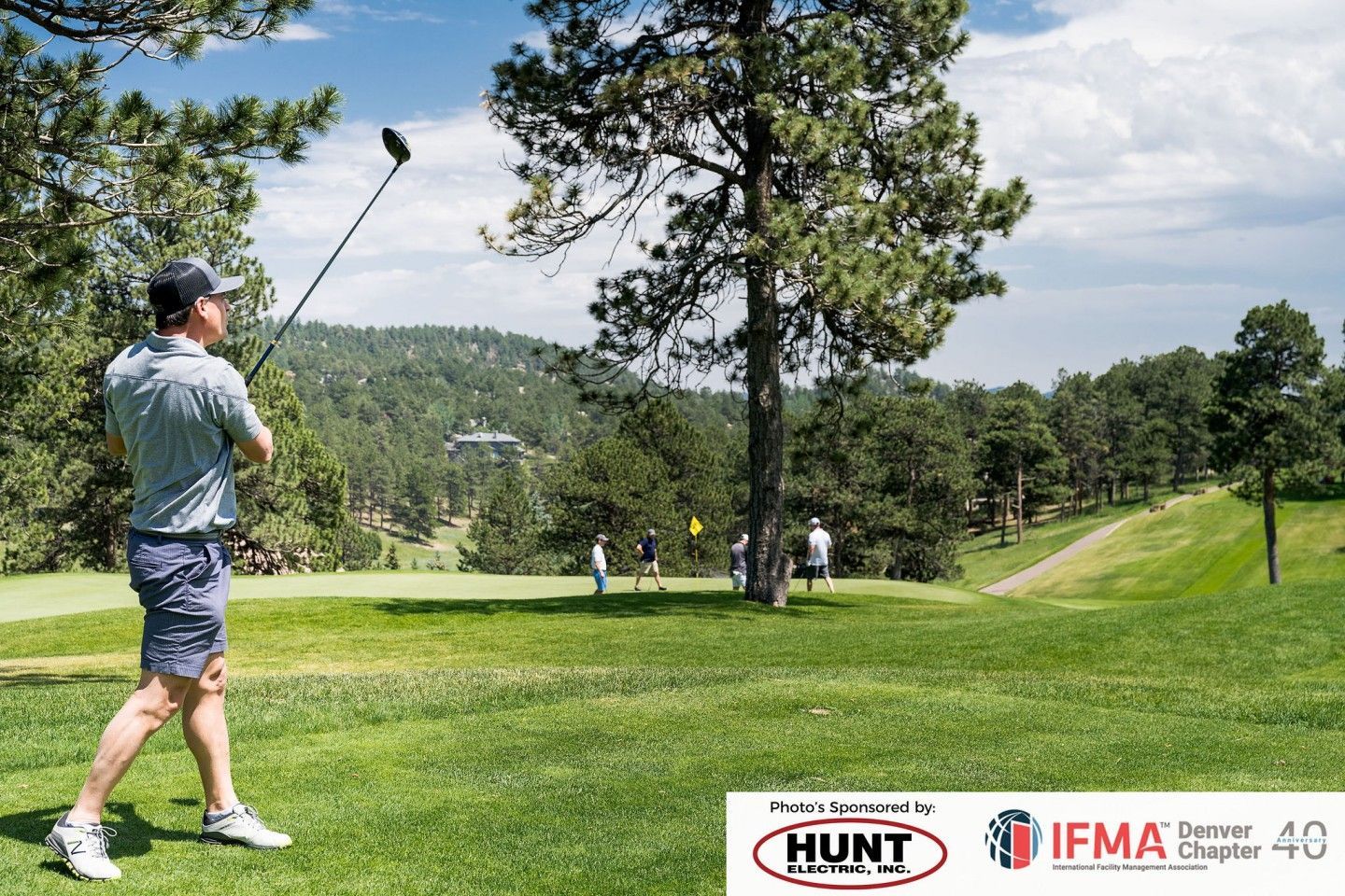 Golfer swings club on green course with trees, hills, and other golfers in the background on a sunny day.