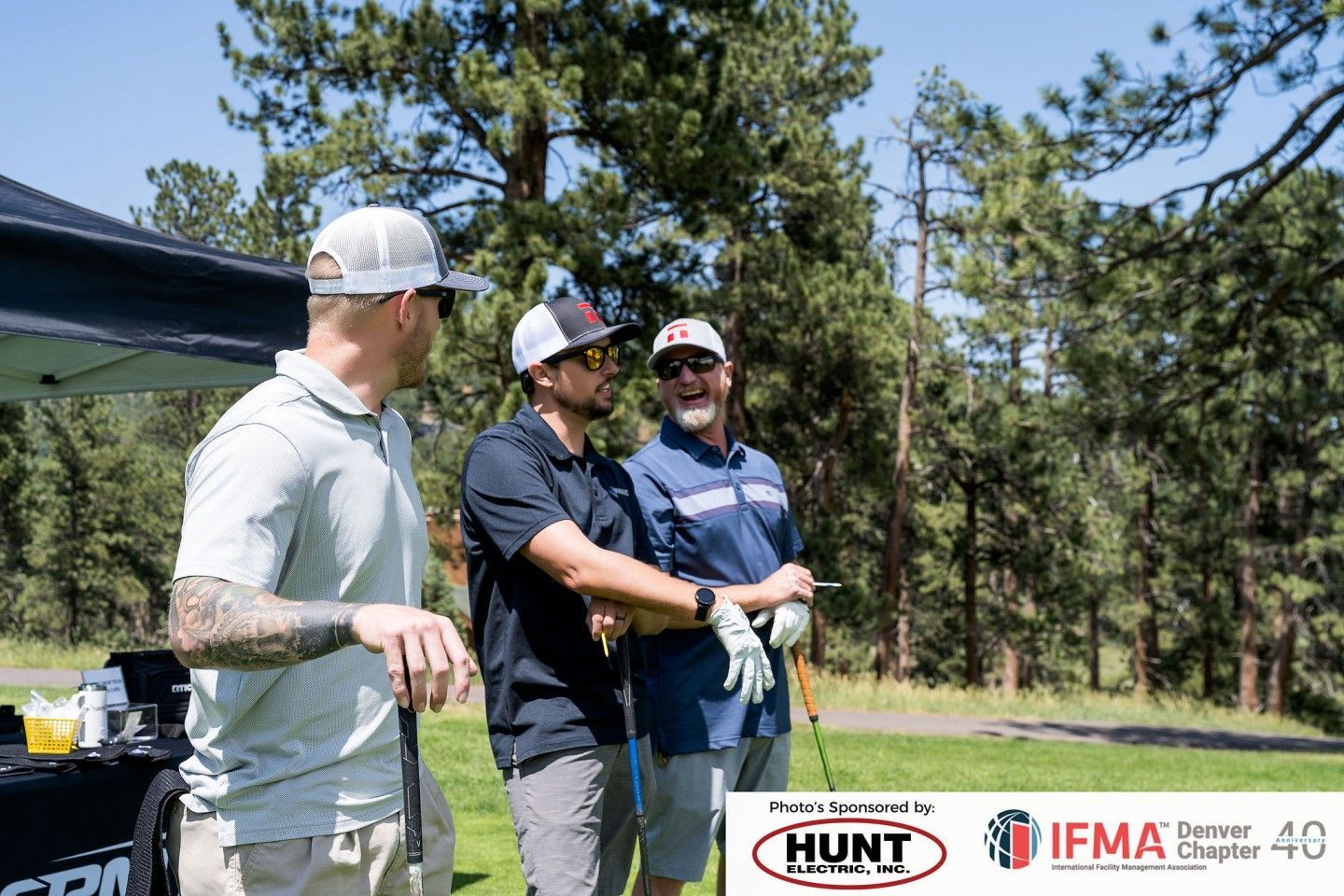 Three men on a golf course laughing, holding clubs. Trees in the background. Sponsors' logos at bottom.