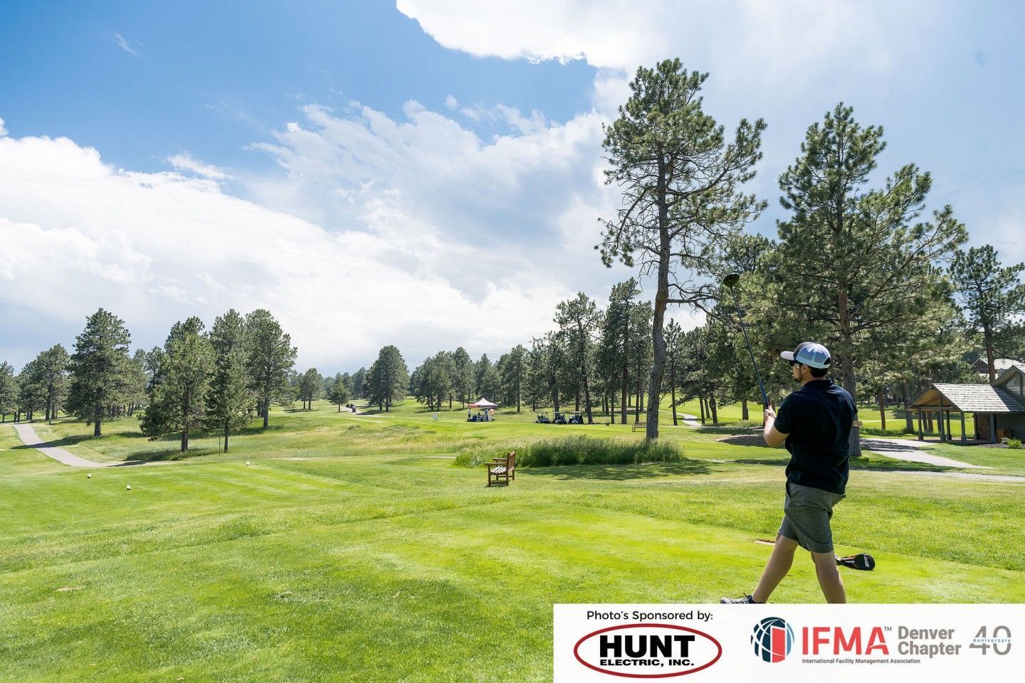 Golfer teeing off on a sunny golf course, surrounded by trees under a blue sky.