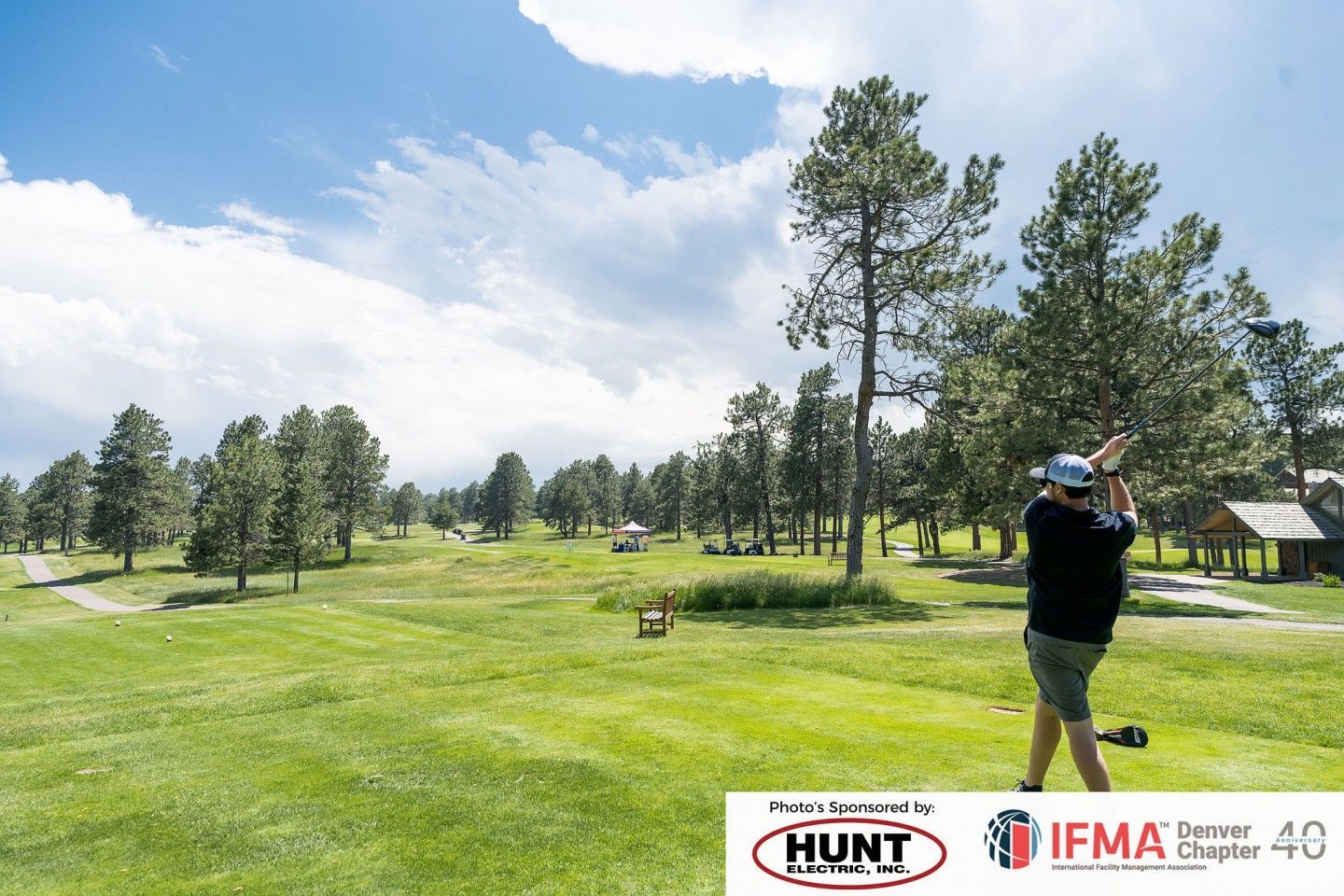 Golfer swinging club on a green course, trees in the background, sunny sky.