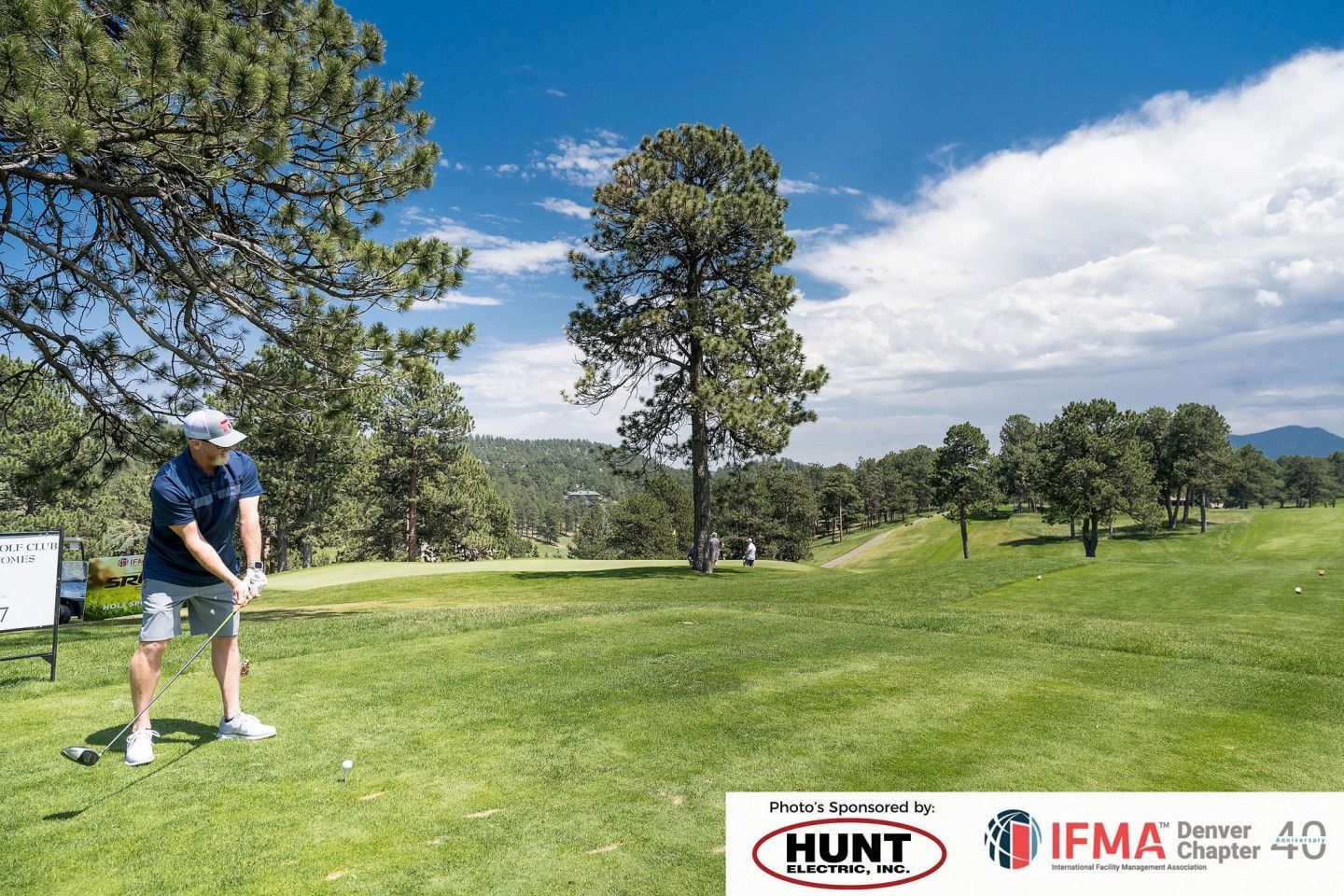 Man swings a golf club on a green course, blue sky and trees in the background.