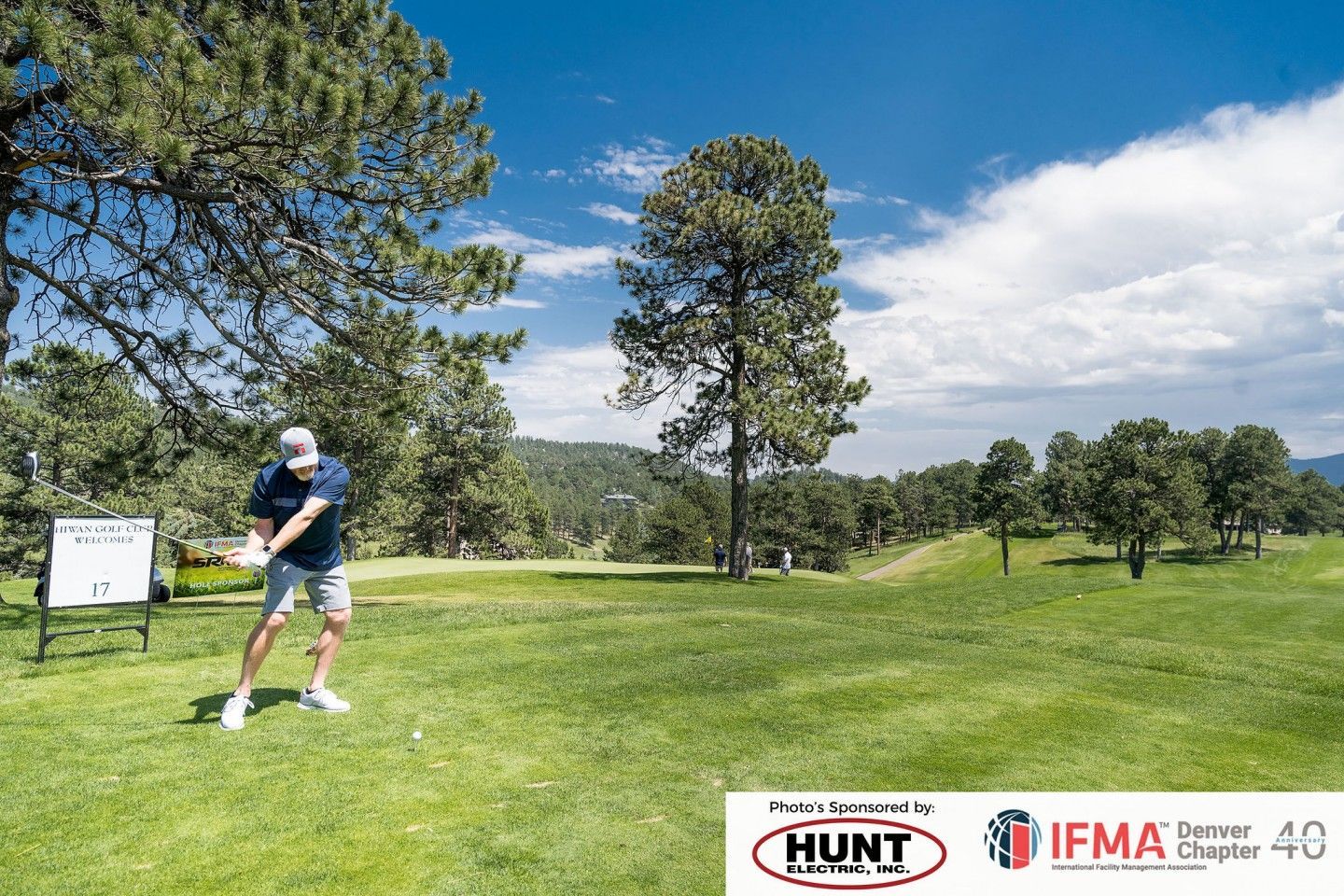 Golfer swings club on green course with trees under blue sky.