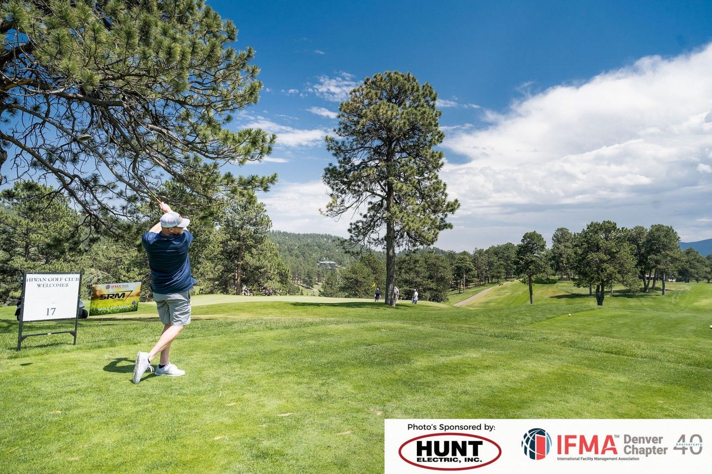 Golfer swinging club on a green course under a blue sky, trees in the background.