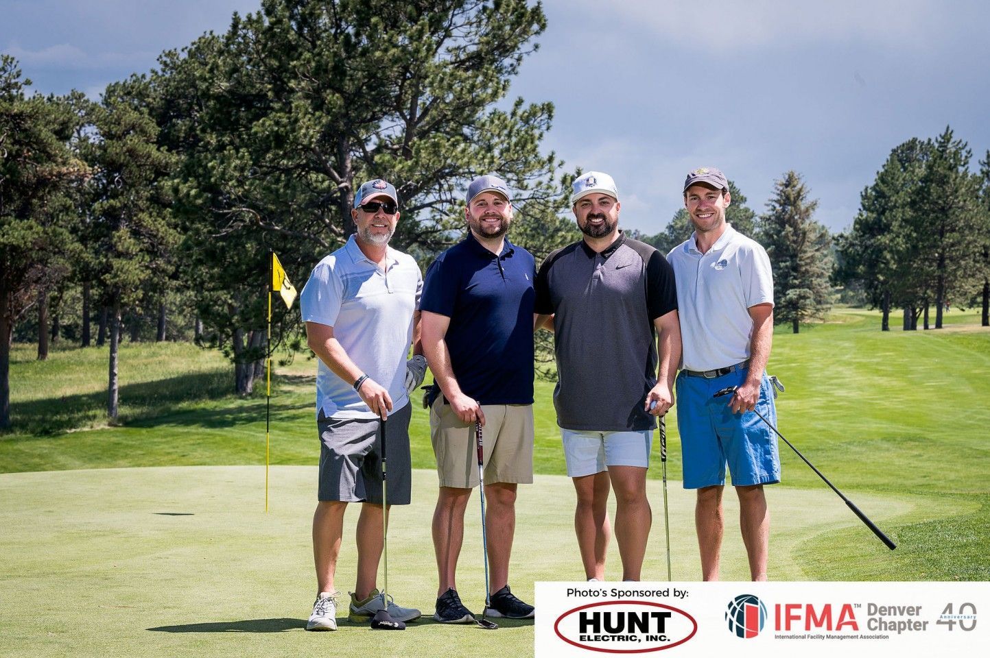 Four men smiling on a golf course, holding clubs. Green grass, trees, and sunny day.