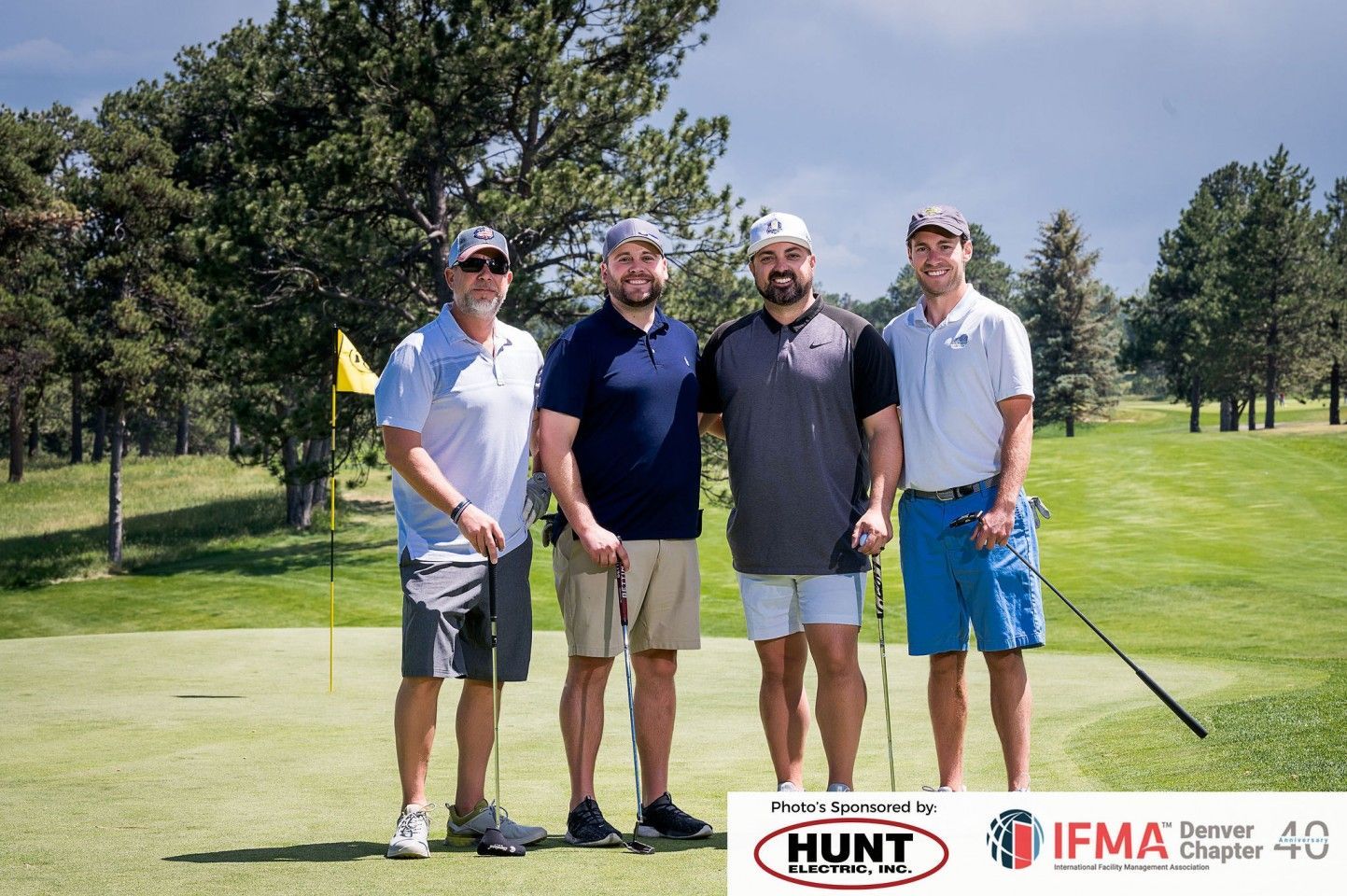Four men on a golf course posing near a green. They are holding golf clubs. Sunny day.