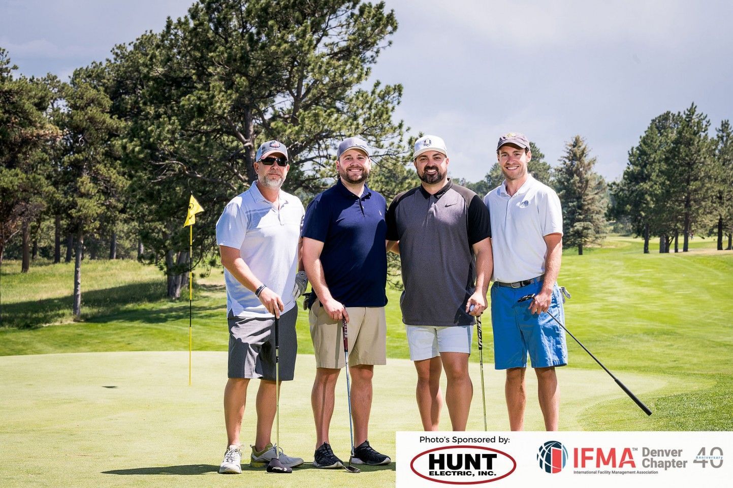 Four men on a golf course pose near a green. Two have their arms around each other.