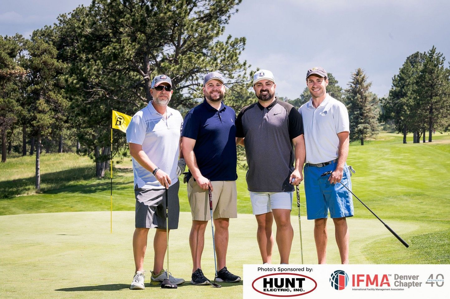 Four men pose on a golf course green. They are wearing golf attire, holding clubs, and smiling. Logos appear at the bottom.