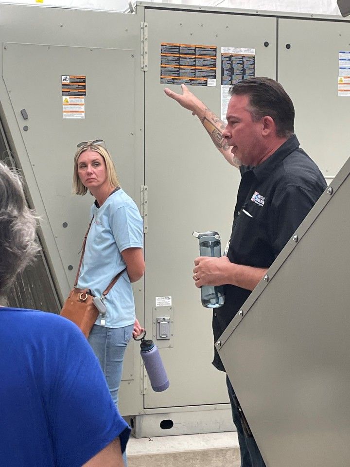 Man pointing at HVAC unit, holding water bottle, talking to a group. Woman in background looks on. Outdoor setting.