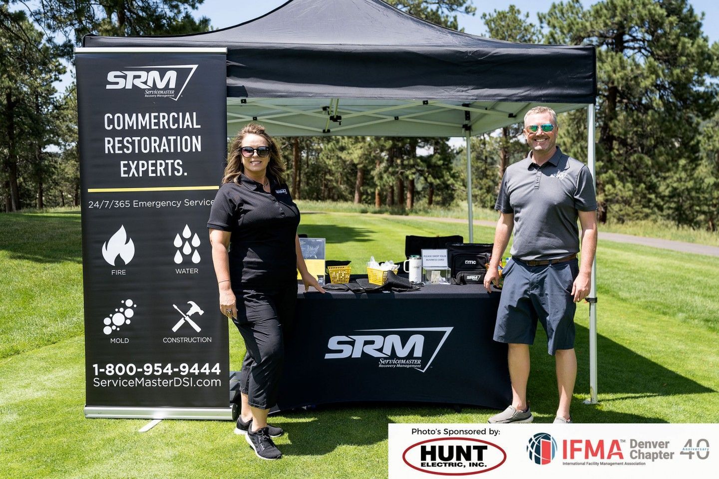 Two people at an outdoor SRM Company booth. Woman on left, man on right. Banner says 