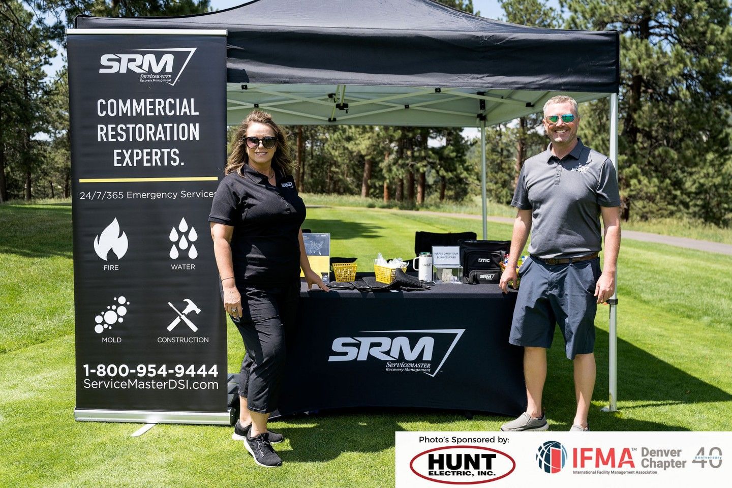 Two people at a SRM commercial restoration booth at an outdoor event. Banner and table display company logo.