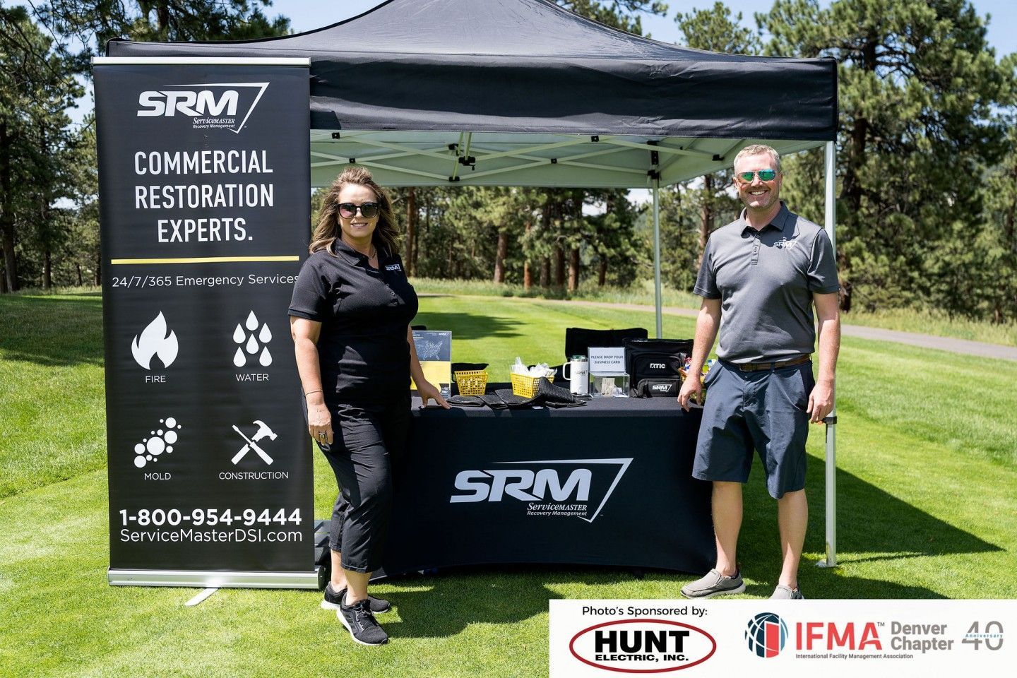 Two people at an SRM Restoration tent at an outdoor event. A banner highlights services with a phone number.