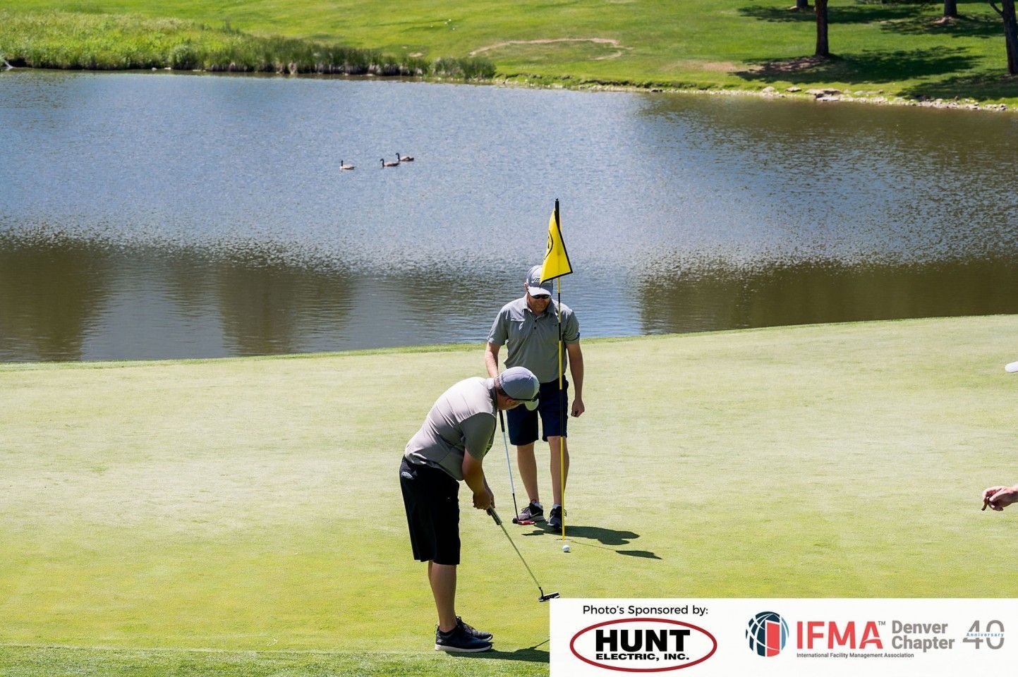 Two men playing golf on a green, near a lake. One is putting, the other watches.