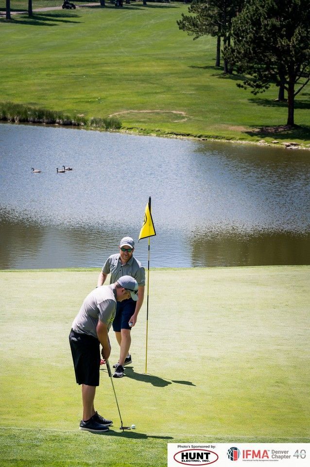 Two golfers on a green, one putting while the other watches near a flag on a lakeside course.