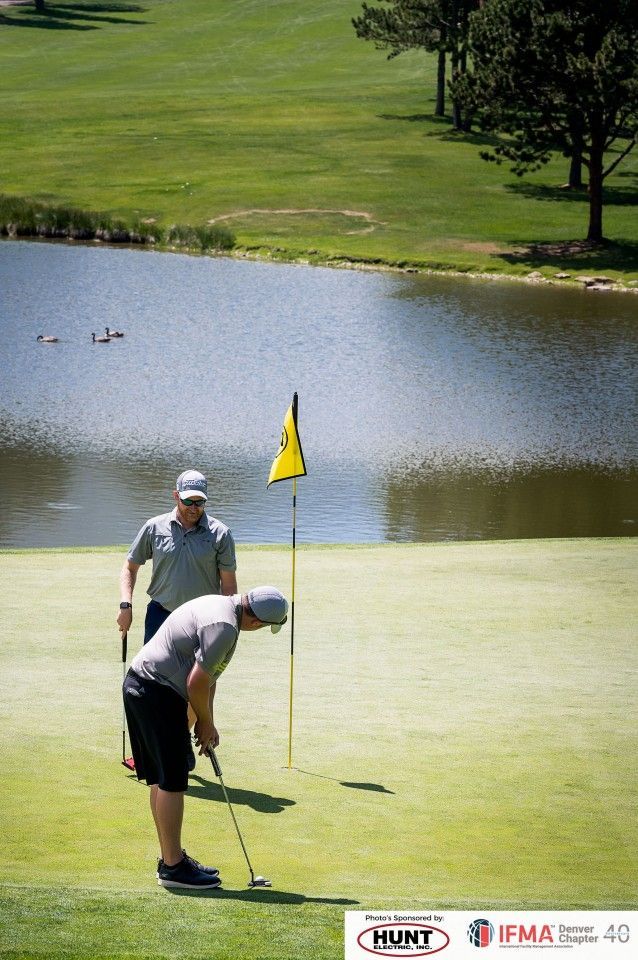Two men golfing near a pond. One putts, other watches. Green grass, yellow flag, sunny day.