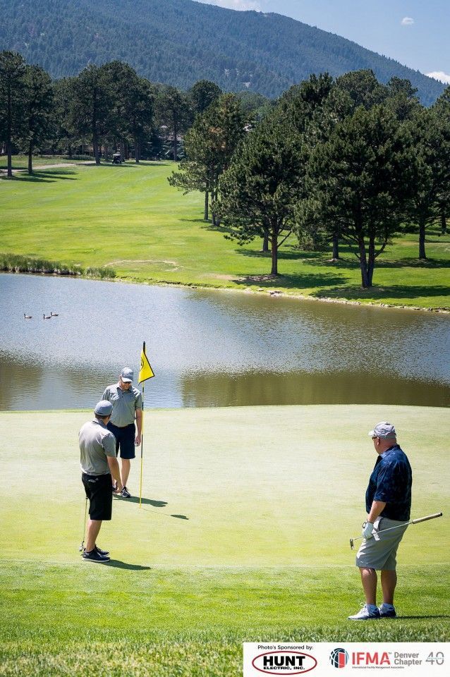 Golfers on a green near a pond; one putts while two watch. Trees and mountains in the background.