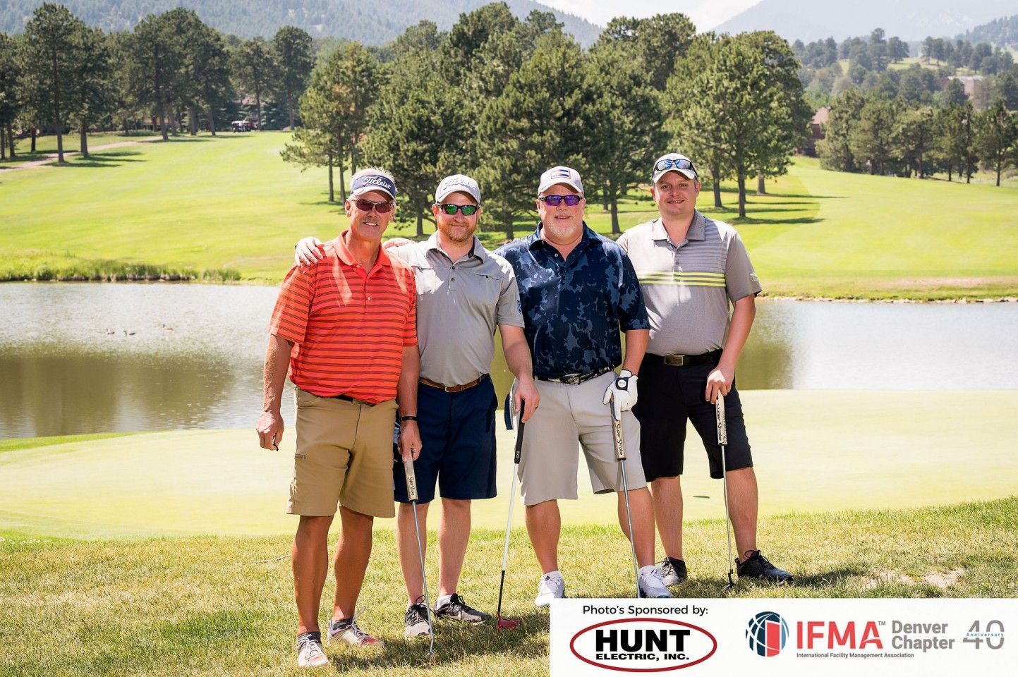 Four men in golf attire pose on a golf course near a lake, smiling.