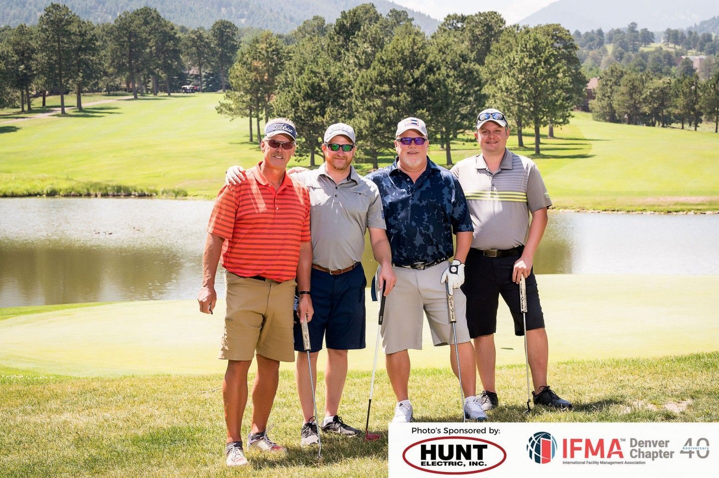 Four men posing on a golf course. They are wearing golf attire, holding clubs. Lake and trees in the background.