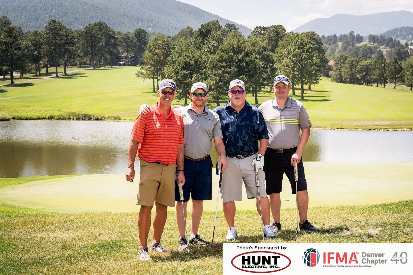 Four men posing on a golf course, mountains in the background, near a lake.