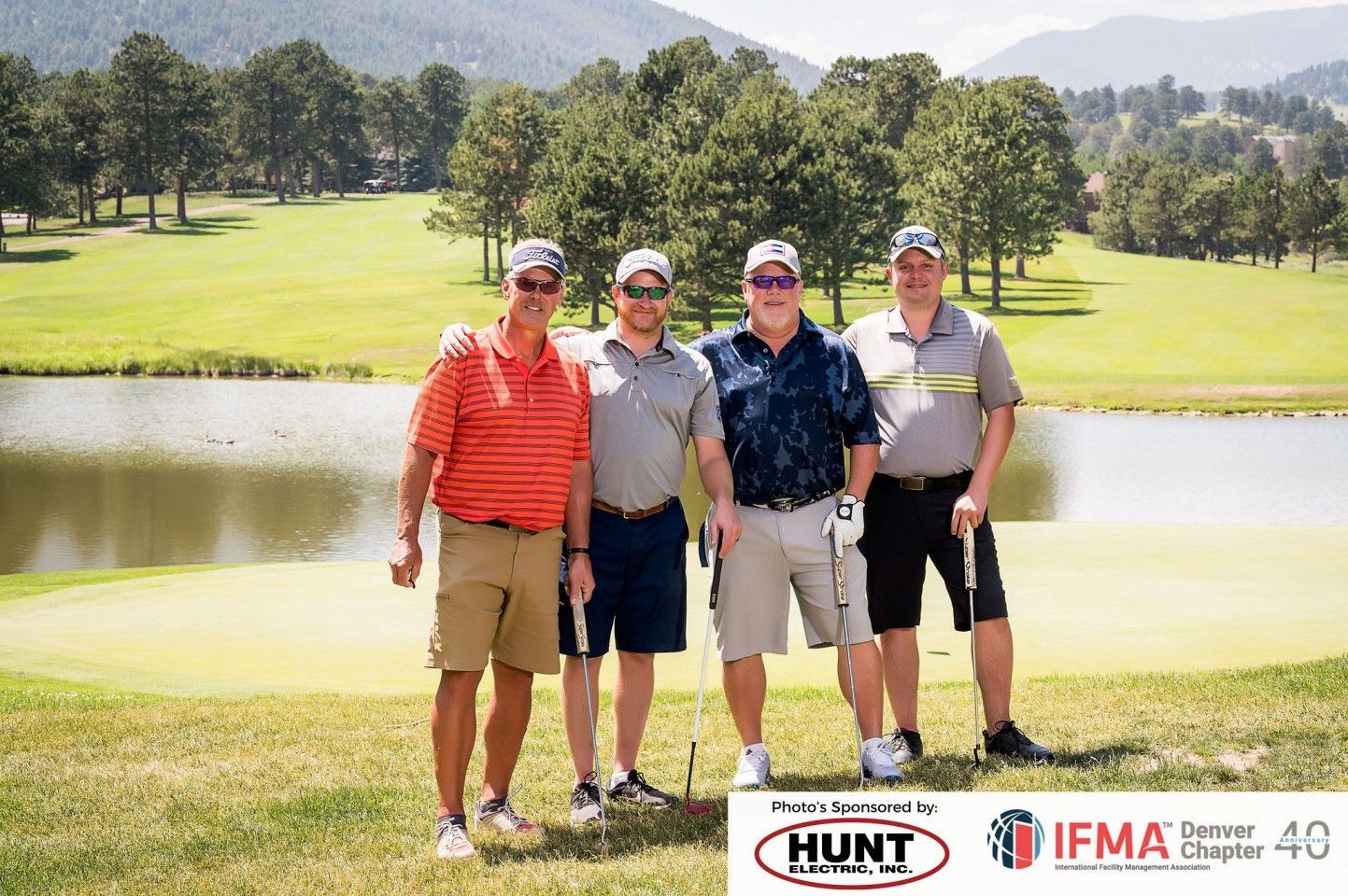 Four men posing on a golf course. They wear casual attire, holding golf clubs near a lake with a mountain backdrop.