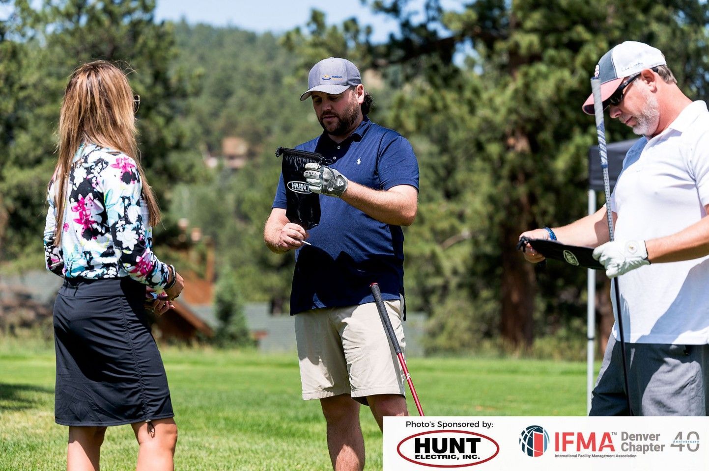 Three people on a golf course. One woman and two men. The men are holding golf clubs and gear. Green grass, trees.