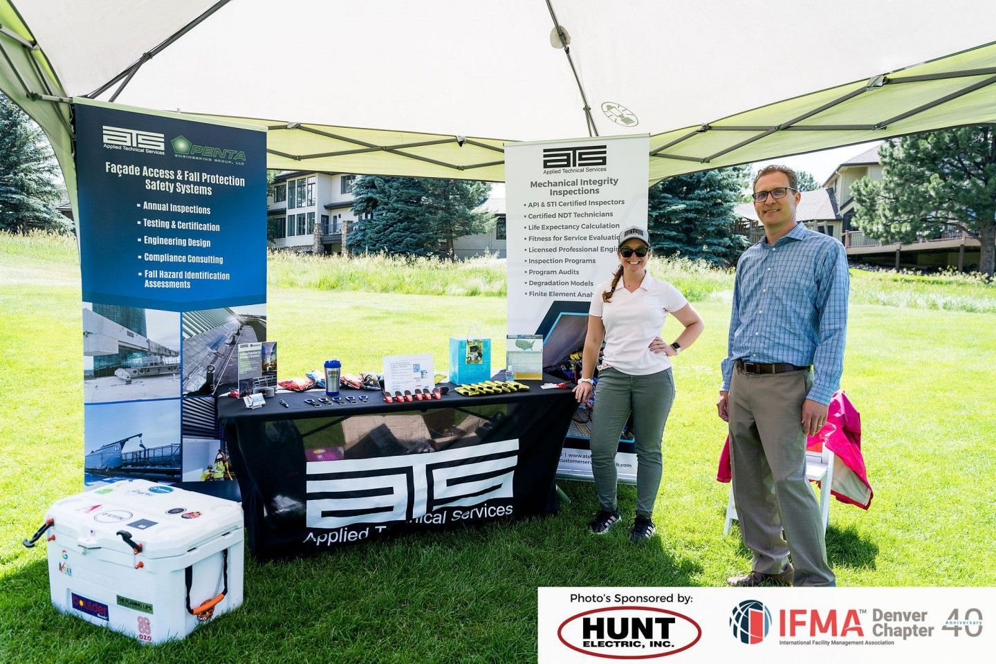 Two people stand at a booth under a tent on grass. Banner with company logo and products displayed on table.