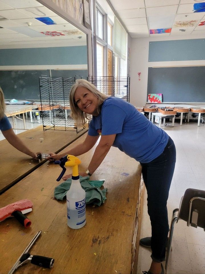 Woman cleaning a wooden table with a spray bottle and cloth in a classroom.
