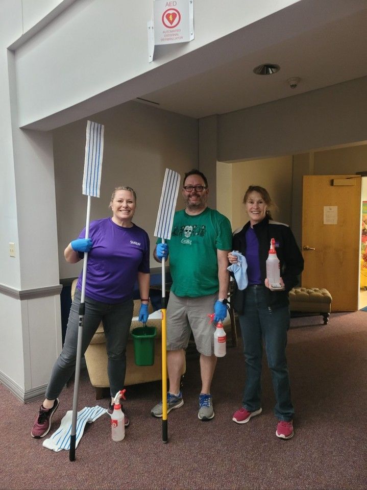 Three people holding cleaning supplies in a hallway: a mop, spray bottles, and rags.