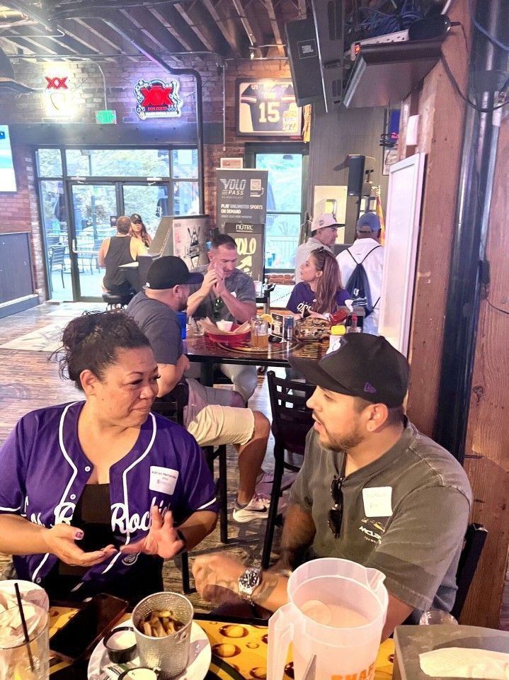 People seated at tables in a bar, conversing and eating. Purple and black clothing, dim lighting.