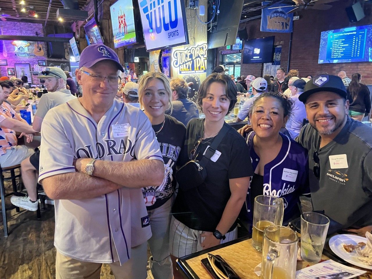 Group of people smiling at a bar, wearing baseball jerseys and hats.