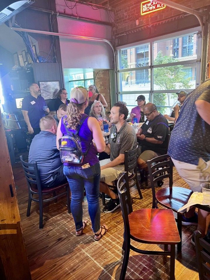 People at tables and standing in a bar, chatting. Open garage door-style windows let in light. Neon sign.