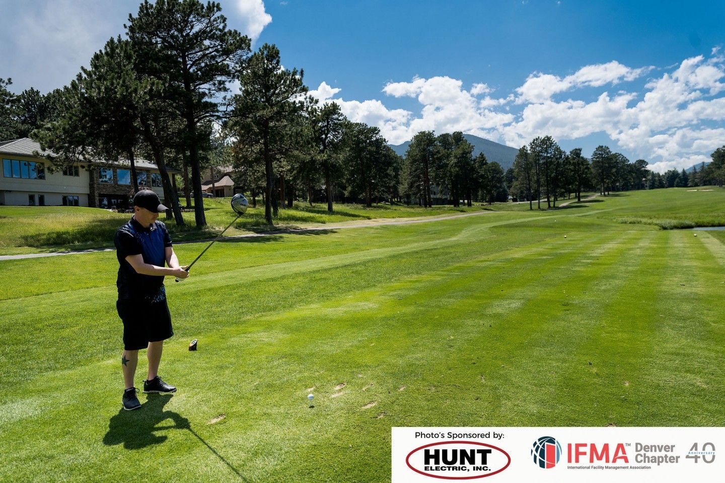 Man in black shirt and shorts preparing to swing a golf club on a green course, blue sky, trees